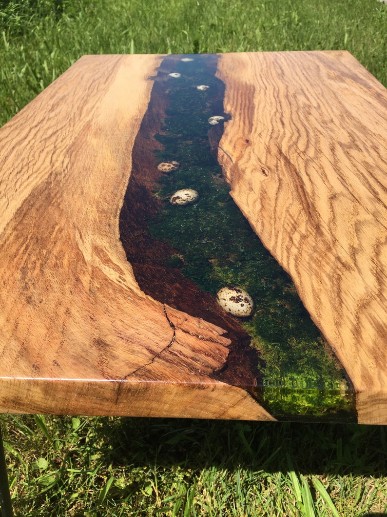 Pode incluir: Uma mesa de centro de madeira com um design de borda viva. O tampo da mesa apresenta um rio de resina com um esquema de cores verde e marrom e ovos de codorniz salpicados de branco embutidos na resina.