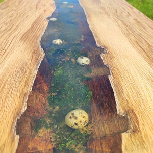 Pode incluir: Uma mesa de madeira com um rio de resina ep&oacute;xi transparente que corre pelo centro. O rio cont&eacute;m musgo e v&aacute;rios ovos pequenos brancos.