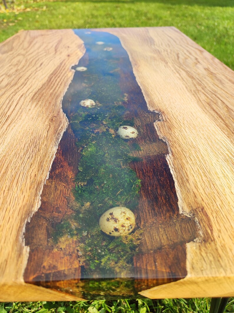 Pode incluir: Uma mesa de madeira com um rio de resina ep&oacute;xi transparente que corre pelo centro. O rio cont&eacute;m musgo e v&aacute;rios ovos pequenos brancos.