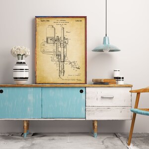 Pode incluir: Uma impressão emoldurada de patente vintage de uma máquina, exibida em um armário azul e branco desgastado. A impressão tem um fundo bege com desenhos técnicos e texto em preto. Um vaso com flores brancas e um vaso listrado estão no armário.