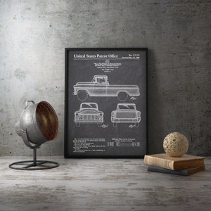 May include: Framed patent illustration of a vintage pickup truck. The black and white drawing shows side, front, and rear views. The frame is black, and the background is a textured grey. A decorative lamp, a ball of twine, and books are on the table.