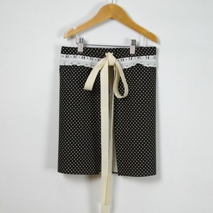 Op de afbeelding: Een zwart-witte stippen schort met een witte kantversiering en een crèmekleurige strik.