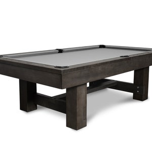 May include: A dark wood pool table with a gray felt top and a simple, modern design. The table has a rectangular shape and a set of four legs.