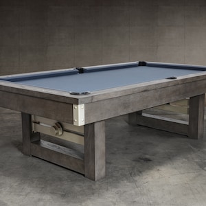 May include: A dark gray wooden pool table with a blue felt top and metal accents. The table has a unique, industrial-style design with exposed beams and a metal support system.