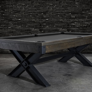 May include: A dark wood pool table with a black felt top and a black metal X-shaped base. The table is set against a brick wall.