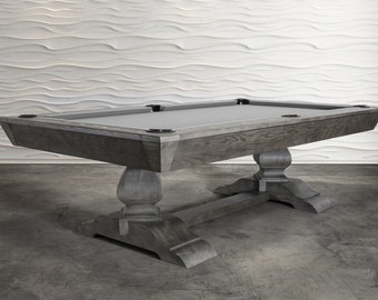 May include: A gray pool table with a gray felt top and a wooden base with four legs. The table is in a room with a white wall.
