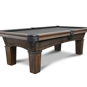 May include: A dark brown wooden pool table with black felt and black metal corner pockets. The table has a traditional design with curved legs and a slightly distressed finish.
