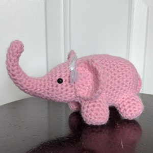 May include: A pink crocheted elephant toy with a white bow on its head. The elephant has a long trunk and small, black eyes.
