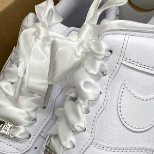 May include: Close-up of white sneakers with satin ribbon laces. The sneakers have a classic design with a perforated toe box and a stitched Nike swoosh. The laces are adorned with silver AF charms.