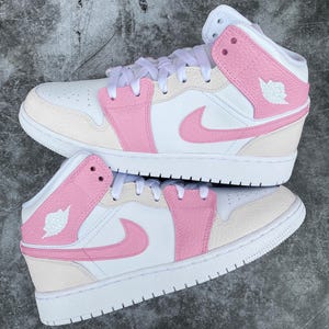 Puede incluir: Un par de zapatillas altas blancas y rosas. Los zapatos presentan una base blanca con detalles en rosa en los paneles laterales, el swoosh y el cuello del tobillo. El logotipo de Jumpman es visible en el lateral. Cordones blancos y suela blanca.