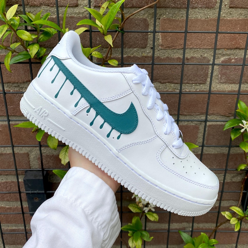 May include: White leather sneaker with teal green accents. The Nike swoosh and a drip design are featured on the side. The word "AIR" is visible on the midsole. White laces complete the look.