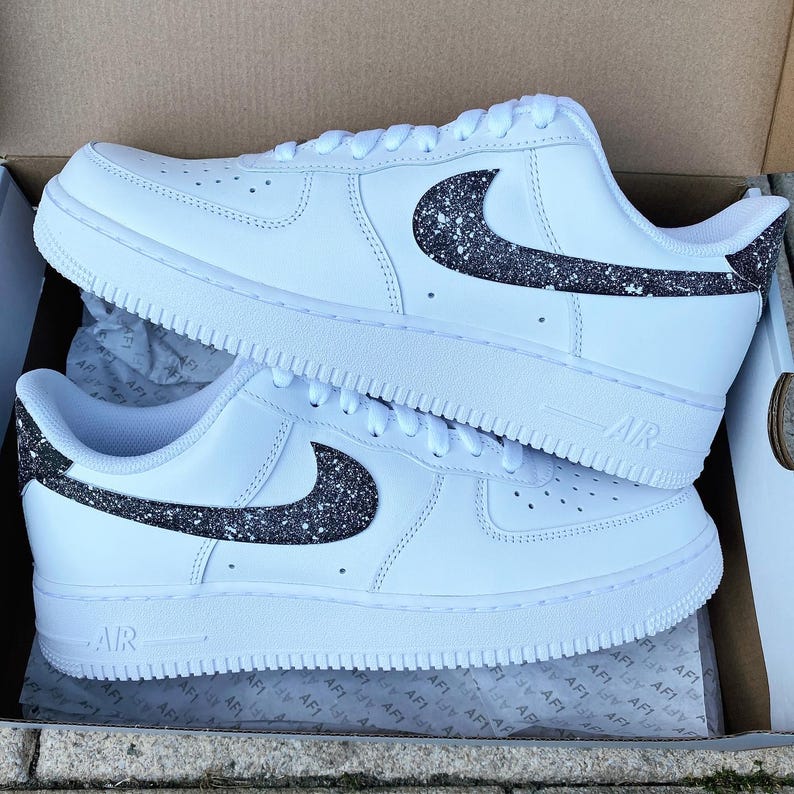May include: White sneakers with a black glitter design on the side and heel. The shoes have a classic design with the word "AIR" visible on the side. The sneakers are in a box.
