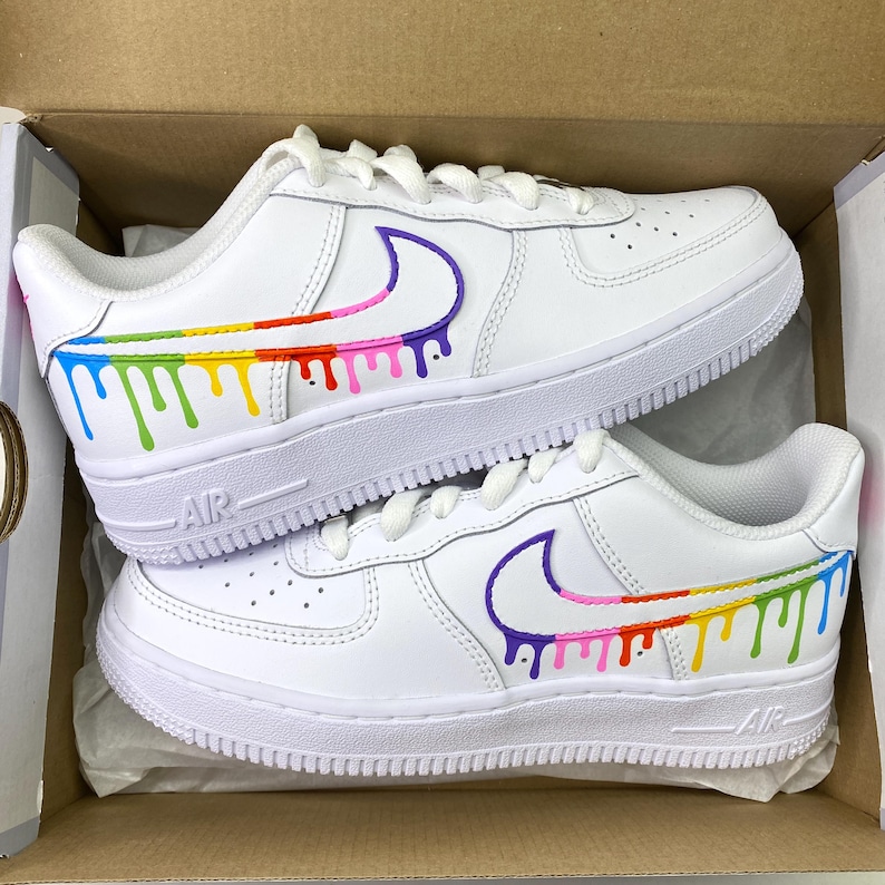 May include: White sneakers with a rainbow drip design. The shoes feature a white base with a colorful drip pattern along the sides and a purple Nike swoosh. The soles are white, and the shoes are in a cardboard box.