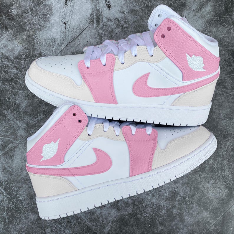 Puede incluir: Un par de zapatillas altas blancas y rosas. Los zapatos presentan una base blanca con detalles en rosa en los paneles laterales, el swoosh y el cuello del tobillo. El logotipo de Jumpman es visible en el lateral. Cordones blancos y suela blanca.