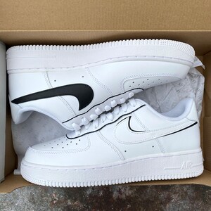 May include: White leather sneakers with black swoosh logos, laces, and a white sole. The sneakers are in a cardboard box, with one shoe on top of the other.