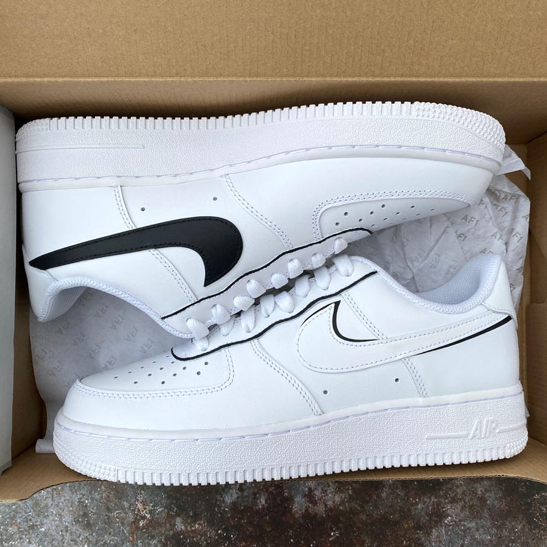 May include: White leather sneakers with black swoosh logos, laces, and a white sole. The sneakers are in a cardboard box, with one shoe on top of the other.