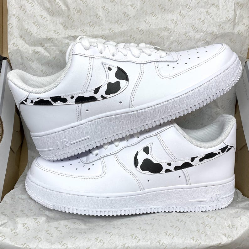 May include: White leather sneakers with black cow print accents on the side and heel. The shoes have a white sole and the word "AIR" is printed on the side. The sneakers are in a box.