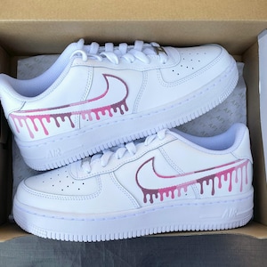 May include: Pair of white leather sneakers with pink and purple drip design on the side. The sneakers have white laces and soles. The word "AIR" is visible on the side of the sole.