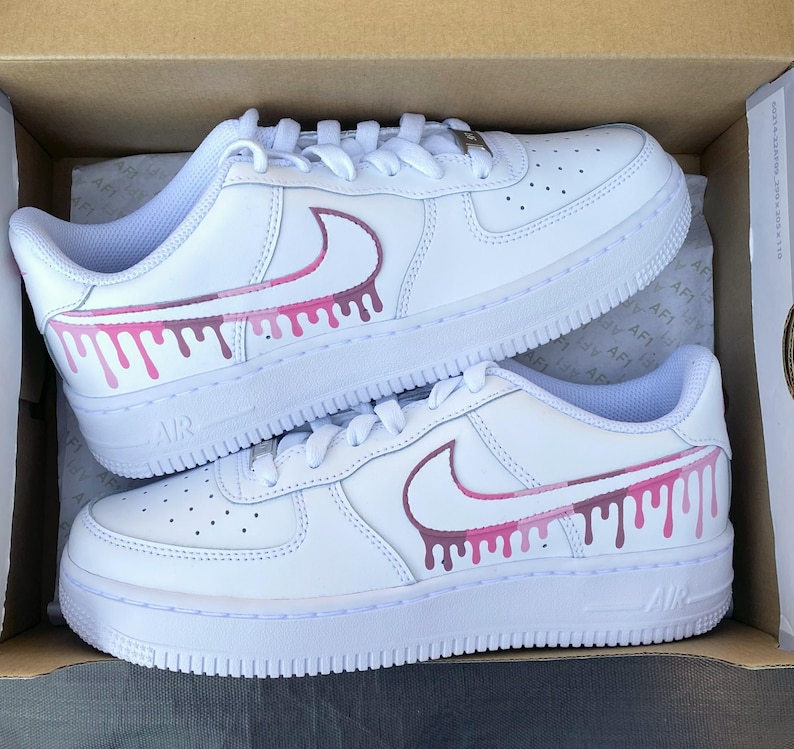 May include: Pair of white leather sneakers with pink and purple drip design on the side. The sneakers have white laces and soles. The word "AIR" is visible on the side of the sole.