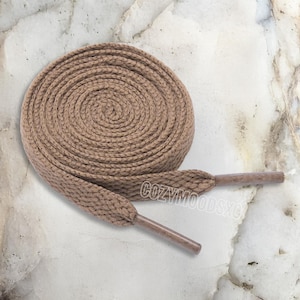 May include: A coiled pair of tan shoelaces with brown aglets. The laces are thick and braided, and the aglets are a solid brown color. The shoelaces are on a light-colored, marbled surface.