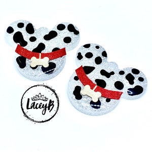 May include: Two Dalmatian-themed Mickey Mouse ear-shaped ornaments. Each ornament has a white base with black spots, a red collar, and a bone-shaped charm. The ornaments are glittery and set against a white background.