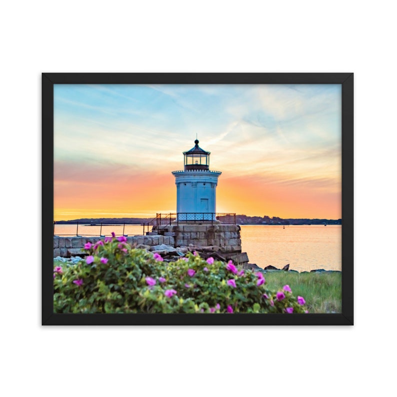 Pu&ograve; includere: Un faro bianco con tetto nero si erge su un molo roccioso con fiori rosa in primo piano. Il cielo &egrave; un mix vibrante di rosa, arancione e blu, creando una bellissima scena di tramonto.