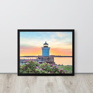 Pu&ograve; includere: Una fotografia incorniciata di un faro bianco su un'isola rocciosa con fiori rosa in primo piano. Il cielo &egrave; di un arancione e rosa vibrante, creando un bellissimo tramonto.