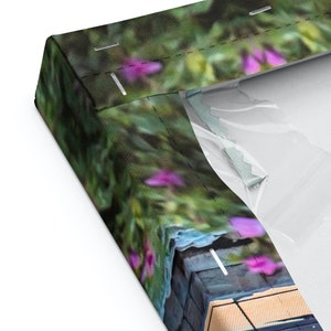 Pu&ograve; includere: Primo piano di una stampa su tela con uno sfondo sfocato di fogliame verde e fiori viola. La tela &egrave; avvolta attorno a una cornice di legno e ha una superficie testurizzata.