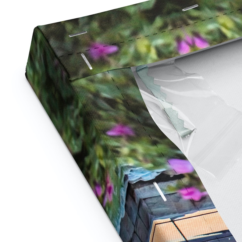 Pu&ograve; includere: Primo piano di una stampa su tela con uno sfondo sfocato di fogliame verde e fiori viola. La tela &egrave; avvolta attorno a una cornice di legno e ha una superficie testurizzata.