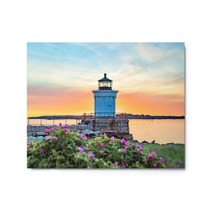 Pu&ograve; includere: Un faro bianco con tetto nero si erge su un molo roccioso, circondato da fiori rosa. Il cielo dietro il faro &egrave; un mix vibrante di rosa, arancione e blu, creando una bellissima scena di tramonto.