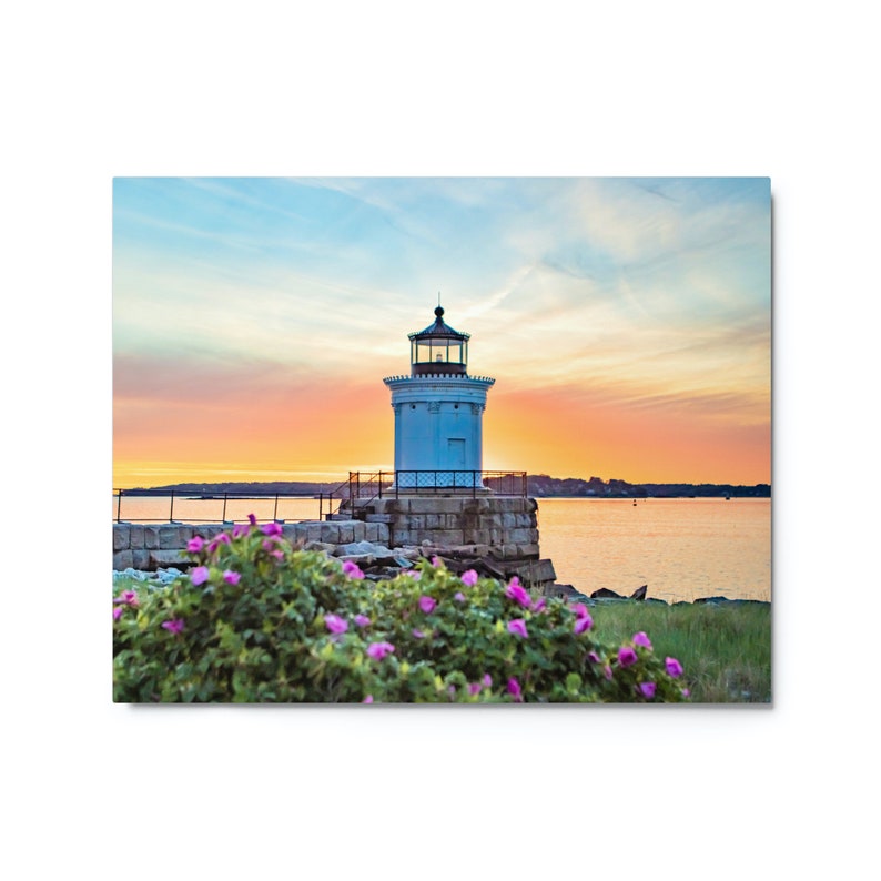 Pu&ograve; includere: Un faro bianco con tetto nero si erge su un molo roccioso, circondato da fiori rosa. Il cielo dietro il faro &egrave; un mix vibrante di rosa, arancione e blu, creando una bellissima scena di tramonto.