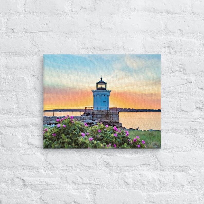 Pu&ograve; includere: Un faro bianco con tetto nero si erge su uno sperone roccioso che domina un'area d'acqua calma. Il cielo &egrave; un mix vibrante di rosa, arancione e blu, creando una bellissima scena di tramonto. I fiori rosa sbocciano in primo piano.