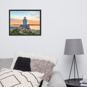 Pu&ograve; includere: Un faro bianco con tetto nero si erge su uno sperone roccioso con fiori rosa in primo piano. Il faro &egrave; in silhouette contro un cielo di tramonto colorato sopra un'area d'acqua.