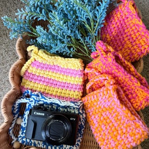 Puede incluir: Una colección de pequeñas bolsas de ganchillo hechas a mano en tonos vibrantes de rosa, naranja y amarillo, junto a una bolsa de rayas azules y blancas que contiene una cámara digital Canon G7X negra, todo en una cesta de mimbre.