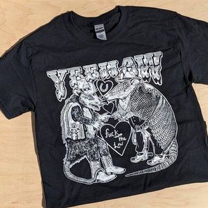 May include: Black t-shirt with a white graphic featuring a possum and an armadillo in cowboy attire, shaking hands. The text "YEEHAW" is above them, with "fuck the law" on a heart below.