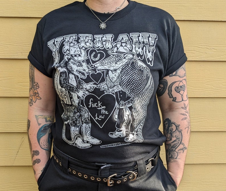May include: Black t-shirt with a white graphic featuring a rat and an armadillo in cowboy attire, shaking hands. The text "YEEHAW" is above them, and "fuck the law" is inside a heart below. The shirt has rolled-up sleeves.