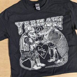 May include: Black t-shirt with a white graphic featuring a rat and an armadillo in cowboy attire, with the text "YEEHAW" above and "fuck the law" on a heart. The design includes a horseshoe and hearts.