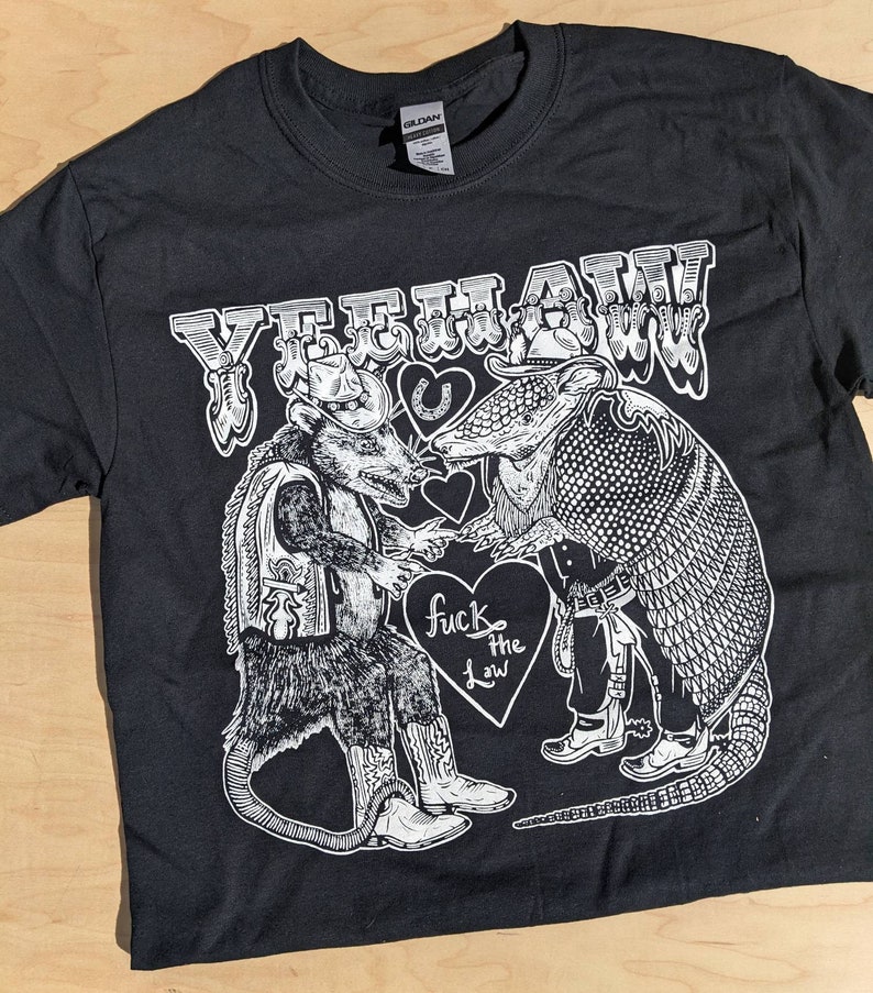 May include: Black t-shirt with a white graphic featuring a rat and an armadillo in cowboy attire, with the text "YEEHAW" above and "fuck the law" on a heart. The design includes a horseshoe and hearts.