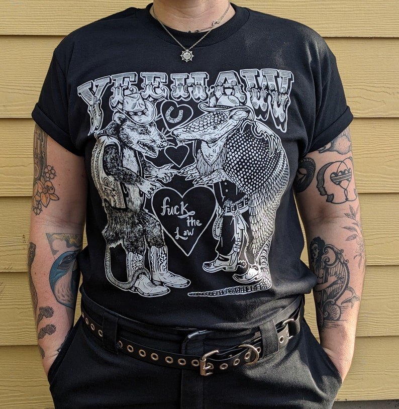 May include: Black t-shirt with a graphic featuring a rat and an armadillo in cowboy attire, the text "YEEHAW!" across the top, and a heart with "fuck the law" in the center. The shirt has rolled-up sleeves.