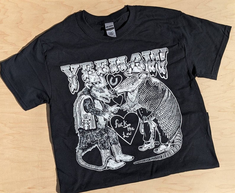 May include: Black t-shirt with a white graphic featuring a possum and an armadillo in cowboy attire, shaking hands. The text "YEEHAW" is above them, with "fuck the law" on a heart below.