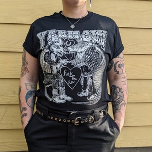 May include: Black t-shirt with the word "YEEHAW!" above a graphic of a rat and an armadillo shaking hands. The graphic includes the text "fuck the law" inside a heart. The shirt has rolled-up sleeves.