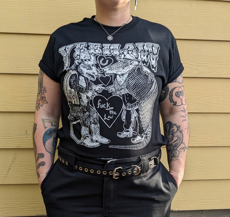 May include: Black t-shirt with the word "YEEHAW!" above a graphic of a rat and an armadillo shaking hands. The graphic includes the text "fuck the law" inside a heart. The shirt has rolled-up sleeves.