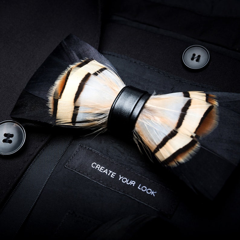 May include: A black bow tie with brown and white feathers and a black leather strap. The bow tie is on a black fabric background with the text "Create Your Look".