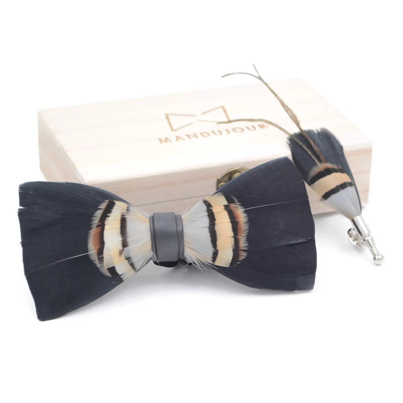 May include: A black and white feather bow tie with a brown accent, in a wooden box with the logo "MANDUJOUR".