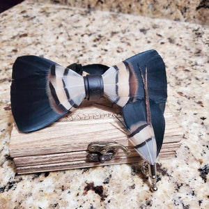 May include: A black and white feather bow tie with a black leather strap and a matching feather pin in a wooden box. The box has the word "Manditio" engraved on it.