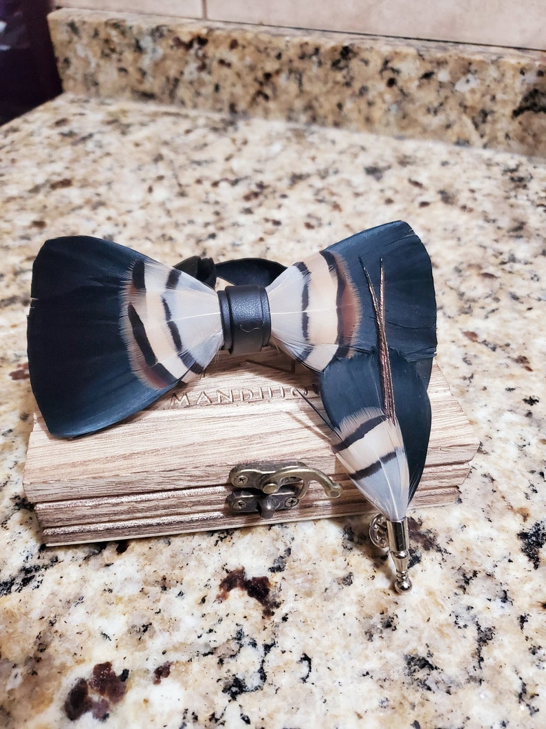May include: A black and white feather bow tie with a black leather strap and a matching feather pin in a wooden box. The box has the word "Manditio" engraved on it.