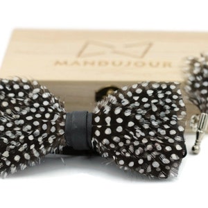 May include: A black and white feather bow tie with a black leather strap. The bow tie is in a wooden box with the word "Mandujour" engraved on the lid.