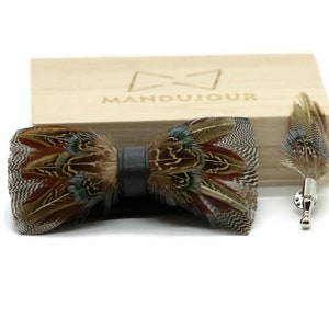 Puede incluir: Una pajarita de plumas de faisán marrón y verde con una correa de cuero negro. La pajarita se muestra en una caja de madera con la palabra "MANDUJOUR" grabada en ella. También se incluye un alfiler de solapa de plumas de faisán a juego.