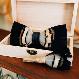 May include: A black bow tie with white and brown feathers, in a wooden box. The bow tie is made of feathers and has a black leather strap.