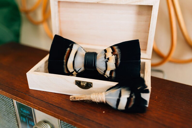 May include: A black bow tie with white and brown feathers, in a wooden box. The bow tie is made of feathers and has a black leather strap.